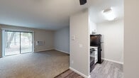 This single-floor unit features a practical, open-concept layout where the living area flows directly into the kitchen. The living space is finished with neutral-toned carpet and painted walls, and is equipped with a wall-mounted air conditioning unit. A sliding glass door provides access to the exterior, allowing natural light into the room. A ceiling fan overhead adds functionality to the space.

The kitchen is efficiently designed in a galley configuration, featuring white shaker-style cabinetry with black hardware, light granite-patterned countertops, wood-look vinyl plank flooring, and black appliances including a refrigerator, range, and dishwasher. Ample upper and lower cabinet storage is present throughout.

The bedroom is a generously sized, carpeted room with neutral gray walls, baseboard heating units, and a window offering views of surrounding greenery. A six-panel door leads to an adjoining area with additional closet access.

The bathroom includes a full-size soaking tub with an integrated white surround, overhead shower, chrome fixtures, and wood-look flooring. The unit maintains a clean, neutral palette throughout, offering a functional and move-in ready living environment.
