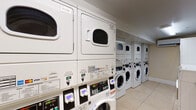 This shared laundry facility is designed to accommodate multiple users efficiently. The room is lined with commercial-grade front-loading washing machines and stacked dryer units along both walls, providing ample capacity for regular use. The machines feature digital payment systems, accepting various payment methods for added convenience. Light tile flooring runs throughout the space, contributing to a clean and functional aesthetic. Overhead fluorescent lighting ensures the room is well-lit. A stainless steel folding table is positioned centrally, offering a practical surface for sorting and folding laundry. Along one wall, a cork bulletin board displays posted notices, laundry tips, and building guidelines in both English and Spanish, keeping users informed. Safety features include a mounted fire extinguisher and a security camera. A wall-mounted mini-split air conditioning unit helps regulate the room's temperature. The entry door includes a window panel, and posted signage throughout the room reinforces proper usage procedures. Overall, the facility is well-organized, clearly maintained, and designed to serve the practical laundry needs of its users with efficiency and accessibility in mind.