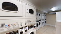 This shared laundry facility is designed to accommodate multiple users efficiently. The room is lined with commercial-grade front-loading washing machines and stacked dryer units along both walls, providing ample capacity for regular use. The machines feature digital payment systems, accepting various payment methods for added convenience. Light tile flooring runs throughout the space, contributing to a clean and functional aesthetic. Overhead fluorescent lighting ensures the room is well-lit. A stainless steel folding table is positioned centrally, offering a practical surface for sorting and folding laundry. Along one wall, a cork bulletin board displays posted notices, laundry tips, and building guidelines in both English and Spanish, keeping users informed. Safety features include a mounted fire extinguisher and a security camera. A wall-mounted mini-split air conditioning unit helps regulate the room's temperature. The entry door includes a window panel, and posted signage throughout the room reinforces proper usage procedures. Overall, the facility is well-organized, clearly maintained, and designed to serve the practical laundry needs of its users with efficiency and accessibility in mind.