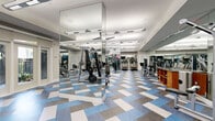 Located at 11070 Katy Freeway in Houston, TX, this fitness center offers a generously sized, single-floor exercise space designed to support a wide range of workout routines. The layout is open and thoughtfully organized, providing ample room to move between equipment without feeling crowded.

The facility is outfitted with a comprehensive selection of cardio machines, including treadmills and stair climbers, as well as a full array of strength training equipment such as cable machines, weight benches, and a dedicated free weights area stocked with dumbbells and medicine balls. A designated TRX Training Zone provides suspension training capabilities, rounding out the facility's diverse fitness offerings.

Mirrored walls line much of the space, enhancing the sense of openness while allowing users to monitor their form. Large mirror columns serve as architectural dividers that define workout zones without obstructing sightlines. Motivational wall graphics add an energetic atmosphere throughout.

Overhead, bright linear lighting ensures the entire floor is evenly and well illuminated. The flooring features a bold geometric pattern in blue, gray, and white, adding a modern, dynamic aesthetic to the space. Wall-mounted flat-screen televisions are positioned throughout for entertainment during workouts. Natural light filters in through windows along one wall, contributing to an inviting and energizing environment.