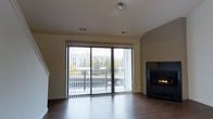 This two-floor space offers a thoughtfully designed layout with clean, modern finishes throughout. The main living area is a standout feature, centered around a sleek gas fireplace with a dark surround that serves as a striking focal point. Large sliding glass doors span a generous portion of the wall, flooding the room with natural light and providing direct access to an outdoor balcony. The balcony overlooks a landscaped neighborhood setting with mature trees and surrounding buildings visible in the background, offering a pleasant outdoor retreat.

The living area is finished with rich, dark hardwood flooring that contrasts beautifully against the warm, neutral-toned walls. Vaulted or angled ceiling details add architectural interest and contribute to the open, airy feel of the space. Vertical blinds on the sliding doors allow for flexible light control and privacy.

Recessed ventilation and ceiling fixtures are neatly integrated into the ceiling, reflecting a well-planned interior design. The staircase, partially visible, connects to the second floor, suggesting additional living or sleeping spaces above. Overall, the space combines functional design with comfortable, inviting elements, making it well-suited for everyday living.