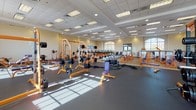 This single-floor fitness facility is designed to accommodate a wide range of training needs in a bright, organized environment. The facility is anchored by a expansive main gym floor finished with dark rubber flooring, housing an impressive array of strength machines, benches, and free weights — all in a cohesive orange and purple color scheme. Large arched windows line the walls, flooding the space with natural light and offering views of the surrounding greenery.

A dedicated cardio room features rows of TRUE-brand treadmills and elliptical machines, each positioned facing wall-mounted flat-screen televisions for an engaging workout experience. Natural light pours in through a prominent arched window at the far end of the room.

A functional training zone provides open floor space outfitted with battle ropes, a heavy tire, cable machines, and a dumbbell rack, ideal for high-intensity and cross-training workouts. Additional strength training areas feature squat racks, barbell stations, and cable pull systems with protective rubber mat flooring.

The facility entrance features a welcoming reception desk with a curved counter, glass double doors, and arched windows that maintain the architectural character throughout. A smaller auxiliary training room with specialized machines and a treadmill rounds out the space, offering a more private setting for focused training sessions.