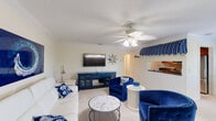 Located at 3124 US Highway 441 SE in Okeechobee, Florida, this single-floor, three-bedroom, one-bathroom home is thoughtfully designed with a cohesive coastal aesthetic throughout. The living and dining area serves as the heart of the home, showcasing plush blue velvet armchairs, a white sectional sofa, a marble-top coffee table, and a decorative palm leaf ceiling fan — all finished with crown molding and a classic blue-and-white nautical theme. A striped awning-style valance frames the pass-through to the kitchen, adding a charming resort-like touch. The kitchen features warm wood cabinetry, dark granite countertops, light hardwood flooring, and stainless steel appliances, offering a functional and well-appointed cooking space. The primary bedroom is a serene retreat with soft lavender walls, plantation shutters allowing ample natural light, and a ceiling fan. A second bedroom continues the coastal motif with rich blue walls, crown molding, plantation shutters, and wall-mounted TV. Three closets, including a spacious walk-in with built-in shelving, provide generous storage. The bathroom features a glass-enclosed walk-in shower with decorative fish tile accents, a vanity, and clean white finishes. A balcony extends the living space outdoors, completing this charming Florida home.