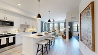 This single-floor model unit in Naples, Florida presents a thoughtfully designed open-concept layout where the kitchen flows seamlessly into the living area. The kitchen is outfitted with white shaker cabinetry featuring black hardware, quartz countertops, stainless steel appliances, a subway tile backsplash, and a central island with seating for casual dining. Pendant lighting and light wood-tone flooring enhance the bright, airy atmosphere throughout.

The living area is generously proportioned, featuring large black-framed windows that invite abundant natural light and outdoor views. A ceiling fan anchors the space, which is styled with a coastal-contemporary aesthetic complemented by neutral tones and layered textures.

Both bedrooms are well-sized, each with multiple windows, ceiling fans, and a calm, restful ambiance. The primary bedroom connects to a spa-inspired bathroom with a dual-sink white vanity, black hardware, a large framed mirror, and a walk-in closet equipped with a custom shelving and drawer system for ample storage. A second bathroom mirrors the same clean, modern finishes. Throughout, light flooring and crisp white walls create a cohesive, welcoming environment.