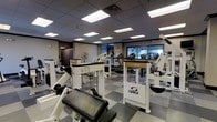 This fitness center is a single-floor facility designed to support a comprehensive workout experience. The space is outfitted with an array of professional-grade Cybex strength-training machines, including cable and resistance units, strategically arranged to allow for comfortable movement between stations. Toward the back of the room, a row of cardio equipment — including treadmills — is positioned in front of large windows that bring in natural light and provide an open, airy feel to the space. The floor features a distinctive checkered tile pattern combining grey and tan tones, adding a clean, structured aesthetic underfoot. Overhead, a dropped ceiling with recessed panel lighting ensures the entire area is consistently and brightly illuminated. A wall-mounted fan provides additional airflow throughout the space. The layout is open and well-organized, offering ample room to navigate between equipment with ease. Overall, the fitness center presents a professional, functional environment suited for a wide range of strength and cardio training activities, making it a well-rounded facility for daily exercise routines.