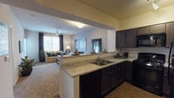 This single-floor residence is designed with an open-concept layout that seamlessly connects the kitchen and living area, creating a cohesive and functional living space. The kitchen is outfitted with dark espresso-toned cabinetry paired with light stone-look countertops, providing a striking contrast. A peninsula with a double-basin sink serves as both a practical workspace and a natural divider between the kitchen and the adjoining living area, enhancing the sense of openness throughout.

The living area features neutral-toned carpeting and is brightened by a window dressed with dark curtains, allowing for adjustable natural light. A ceiling fan adds comfort and practicality to the space. The transition from wood-look flooring in the kitchen to carpeting in the living area subtly delineates the two zones while maintaining visual flow.

Beyond the interior, a private balcony extends the living space outdoors. Finished with textured stucco walls, the balcony offers open views of the surrounding neighborhood, landscaped with mature trees set against a bright, open sky — an inviting spot for outdoor relaxation.