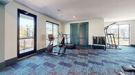 This two-story fitness center is designed to accommodate a wide range of workout needs within a bright, well-appointed space. The ground floor immediately impresses with its soaring double-height ceilings, accented by teal-painted beams and structural columns that define the open layout. Large floor-to-ceiling windows line the perimeter, flooding the space with natural light and offering views of palm trees and outdoor amenities beyond. The lower level is equipped with an extensive selection of strength training equipment, including TKO cable machines, a smith machine, a dedicated Training Zone functional rig with barbells and accessories, and a full dumbbell rack. Kettlebells, plyo boxes, and battle ropes round out the functional fitness offerings, while full-length mirrors enhance the sense of space. A staircase leads to the second floor, which features a more intimate setting with additional cardio machines—including treadmills, ellipticals, and stationary bikes—alongside a dumbbell rack and direct balcony access with outdoor views. Throughout both levels, vibrant patterned carpet in teal and blue tones adds a modern, energetic aesthetic that ties the space together cohesively.
