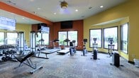 This single-floor fitness center is designed to provide a comprehensive workout experience in a welcoming, well-appointed environment. The space features bold accent walls in warm orange and yellow tones, creating an energetic atmosphere conducive to exercise. Large windows along multiple walls flood the room with natural light while offering outdoor views of surrounding greenery, enhancing the overall sense of openness.

The cardio section is equipped with multiple treadmills and upright stationary bikes, arranged to take full advantage of the natural light and window views. A wall-mounted flat-screen television is positioned centrally, providing entertainment during workouts. A large mirror spans one wall, visually expanding the space and allowing users to monitor their form during strength training.

The ceiling features recessed lighting supplemented by a ceiling fan, ensuring the space remains well-lit and comfortable. The patterned carpet flooring adds texture underfoot while contributing to the room's functional design. Overall, the fitness center balances practicality with an inviting aesthetic, making it a well-rounded facility for a variety of exercise routines.
