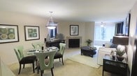 This single-story residence offers an open-concept layout that seamlessly connects the dining and living areas, creating a cohesive and functional living environment. The dining area is anchored by a round glass-top table surrounded by distinctively styled green chairs with sculpted cut-out backs, paired with darker accent chairs, all beneath an elegant pendant light fixture that adds a refined focal point overhead.

The living room flows naturally from the dining space, centered around a corner fireplace with a clean, minimalist surround that serves as an architectural feature of the room. Neutral-toned carpeting throughout lends warmth and continuity across the open floor plan. A dark-finished round coffee table sits atop a light area rug, complementing the living room seating arrangement.

Large windows along the far wall invite generous natural light into the space, enhancing the bright, airy ambiance created by the soft, neutral wall tones. Framed artwork decorates the walls, adding visual interest without overwhelming the understated palette. A console table positioned near the fireplace provides additional display and storage space, contributing to the overall well-appointed character of this inviting residence.