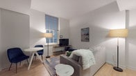 This single-floor unit offers a thoughtfully designed layout that maximizes both functionality and comfort. The open-plan living and dining area features light hardwood flooring, neutral white walls, and notably tall ceilings that lend an airy sense of volume to the space. A large window fitted with slatted blinds draws in natural light, brightening the room. The living area accommodates a plush sofa, a round marble-top coffee table, and a compact dining set with blue accent chairs, creating a cohesive and inviting atmosphere.

The kitchen flows directly from the living space and is equipped with warm wood cabinetry, granite countertops, a white tile backsplash, and a full suite of stainless steel appliances including a range, microwave, and dishwasher, making it both practical and visually appealing.

The bedroom is a serene retreat, featuring plush carpeting, generous closet storage with louvered doors, and two large windows dressed with teal curtains that allow for ample natural light. A wooden dresser and bedside furnishings complete the room.

The full bathroom is finished with neutral tile flooring, white subway-tiled walls, a tub-shower combination, and a sleek wooden vanity, maintaining the clean and contemporary aesthetic found throughout the unit.