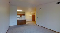 This single-floor unit offers an open-concept design that seamlessly connects the kitchen and living area, creating a spacious and functional layout. The living area is finished with neutral wall-to-wall carpeting, providing a comfortable and cohesive feel throughout the space. Vaulted ceilings add a sense of openness and volume, enhancing the overall airiness of the unit.

The kitchen is positioned adjacent to the living space and features warm medium-toned wooden cabinetry, a white refrigerator, and a white electric range, complemented by under-cabinet lighting that brightens the workspace. A peninsula with a dark wood-paneled base and light countertop serves as a natural divider between the kitchen and living area, offering additional prep space and a casual gathering point.

A small dining area is suggested by the elegant chandelier that hangs centrally between the kitchen and the entryway, casting a warm glow over the transition space. The solid wood entry door adds a traditional touch and connects to a hallway that leads further into the unit. Neutral walls and consistent trim throughout contribute to a clean, move-in-ready aesthetic.