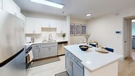 Located at 5005 Community Circle in Charlotte, NC, this 2-bedroom, 2-bathroom single-floor residence offers a thoughtfully designed layout that maximizes comfort and functionality. Light wood-tone flooring flows continuously throughout, complementing the clean white walls and creating a bright, cohesive aesthetic.

The open-concept living, dining, and kitchen area forms the heart of the home. The living room features large windows and a glass door that flood the space with natural light, while the kitchen stands out with white upper cabinetry, two-toned grey lower cabinets, quartz countertops, stainless steel appliances, and a central island with bar seating — ideal for both meal prep and casual dining.

A central hallway connects the bedrooms and bathrooms, offering a clear and efficient flow. The primary bedroom is a comfortable retreat with a glass door to the exterior, generous natural light, and ample closet space. The second bedroom is equally well-appointed. Both bathrooms feature grey shaker-style vanities with white quartz countertops, modern light fixtures, and large mirrors, reflecting a consistent and polished design language.

With four closets distributed throughout, storage is plentiful. The overall ambiance is modern, clean, and welcoming, making this Charlotte residence a practical and stylish place to call home.