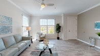 This single-floor residence is thoughtfully designed with a clean, contemporary aesthetic carried throughout by a neutral color palette of soft grays, warm taupes, and crisp whites. The living room serves as the heart of the home, featuring light wood-look tile flooring, crown molding, and a ceiling fan overhead. Multiple windows fitted with horizontal blinds flood the space with natural light and offer views of the surrounding greenery. The open layout flows naturally from the living area toward the kitchen and entry.

The kitchen is well-appointed with white shaker-style cabinetry, a mosaic tile backsplash, white quartz countertops, and stainless steel appliances including a refrigerator, range, and over-the-range microwave. A peninsula counter with pendant lighting provides bar-height seating for two, making it an ideal casual dining spot.

Both bedrooms are carpeted and bright, each featuring windows with outdoor views and ceiling fans. The primary bedroom benefits from multiple windows and direct access to an adjoining room. The bathroom includes a wide vanity with white cabinetry, quartz countertops, a large mirror, and a tub-shower combination with decorative tile accenting. Large-format tile flooring completes the clean, functional design.