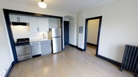 This single-floor space combines classic architectural character with modern finishes, creating a clean and functional living environment. The kitchen is equipped with stainless steel appliances, including a full-size refrigerator and dishwasher, complemented by sleek gray cabinetry and a white subway tile backsplash. A stainless steel sink with a modern faucet adds to the kitchen's polished look. Light-toned hardwood flooring runs throughout, lending warmth and continuity to the space. A standout architectural feature is the elegant arched doorway, which adds a timeless, classic touch and creates a natural visual focal point. Bold black door and window trim provide striking contrast against the light walls, lending the space a modern edge. The layout opens into additional areas, including what appears to be a closet or utility space visible through a secondary doorway, offering practical storage. A ceiling-mounted globe light fixture adds a vintage-inspired charm overhead. The overall ambiance is bright, clean, and thoughtfully designed, balancing period architectural details with contemporary updates for a polished, move-in-ready feel.