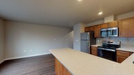 This single-floor residence is thoughtfully designed with an open, functional layout that connects the kitchen and living areas seamlessly. The kitchen is a standout feature, equipped with stainless steel appliances including a refrigerator, electric range, microwave, and dishwasher. Warm-toned wood cabinetry provides ample storage, while a light-colored quartz countertop peninsula extends into the living space, offering additional prep area and casual seating. A tile backsplash adds a subtle decorative element behind the range. Multiple windows fitted with blinds allow natural light to fill the open living and kitchen area, contributing to a bright and welcoming atmosphere. Wood-look plank flooring runs throughout the main living spaces, providing durability and a cohesive aesthetic. The bedroom features plush carpeting and a sliding-door closet with a rod and shelf for organized storage, complemented by recessed lighting overhead. The bathroom is efficiently laid out with clean, neutral finishes, wood-look flooring, and direct access to the adjoining spaces. The overall design palette is neutral and modern, with soft gray and beige tones throughout, creating a clean and move-in-ready environment.