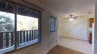 This single-floor space presents a straightforward and functional layout centered around a welcoming entry area. The front door features a classic multi-pane glass insert, allowing natural light to filter into the interior while providing a charming architectural detail. Adjacent to the entry, a generously sized window looks out onto a wooden deck railing and lush, mature tree foliage beyond, bringing the outdoors in and filling the space with abundant natural light.

The entry transitions into an open area finished with light-toned tile flooring, which contributes to a clean and airy feel. A ceiling fan with an integrated light fixture is centrally positioned overhead, offering both air circulation and ambient lighting. Carpeted flooring is present in a portion of the space, adding a layer of texture and warmth to contrast the tile.

Baseboard heating units are visible along the walls, indicating a practical heating system suited to cooler climates. The walls are finished in a neutral white, keeping the interior feeling open and versatile. Overall, the space offers a modest, no-frills environment with functional features and pleasant natural light from the tree-lined outdoor surroundings.