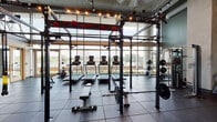 Located at 417 W Peace St in Raleigh, NC, this two-floor fitness center delivers a well-appointed workout experience within a striking industrial-modern setting. Exposed concrete ceilings, large-format ductwork, and visible structural columns define the architectural character throughout, complemented by warm accent lighting and rubber-tiled flooring purpose-built for high-performance training.

The main floor is anchored by a generously sized free weights area equipped with multi-station power racks, barbells, weight plates, dumbbells, and cable machines — all arranged to allow ample workout space. Floor-to-ceiling windows flood the strength training zone with natural light while offering open views of the surrounding Raleigh cityscape.

The cardio zone features rows of treadmills, ellipticals, and a stair climber, each equipped with individual screens. Mirrored walls extend the sense of space, while exposed ceiling infrastructure and ceiling fans maintain an industrial aesthetic. Treadmills are positioned directly along the window line, providing exercisers with engaging outdoor views during workouts.

A modern metal staircase with sleek railings connects the two floors, contributing to the facility's contemporary design language. The upper level provides additional training space overlooking the main floor below. The overall ambiance is energetic yet functional, combining thoughtful equipment placement with architectural details that make this fitness facility both practical and visually compelling.