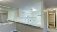 This single-floor space offers a clean and functional layout with a distinctly bright and neutral aesthetic throughout. The kitchen is a standout feature, designed with an open-concept approach and separated from the adjacent living area by a peninsula-style breakfast bar topped with a light speckled countertop — ideal for casual dining or meal prep. The kitchen is outfitted with ample white upper and lower cabinetry, providing generous storage, and is equipped with a white electric range, a matching refrigerator, and an under-cabinet range hood. Overhead, a flush fluorescent light fixture ensures the workspace is well-illuminated. The uniformly white cabinetry and appliances, paired with the light-toned walls and countertops, create a cohesive and airy atmosphere. The neutral color scheme — predominantly whites and soft grays — lends a timeless and versatile quality to the interior, making it easy to personalize. The single-floor plan ensures convenient, step-free living throughout. Overall, the space presents a tidy, move-in ready environment with a straightforward, practical design focused on comfort and functionality.