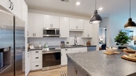 This thoughtfully designed single-floor residence offers a seamless blend of modern style and functional living. The open-concept kitchen is a standout feature, showcasing crisp white shaker cabinetry, a herringbone tile backsplash, dark granite countertops, stainless steel appliances, and a striking navy blue center island topped with matching stone — all illuminated by industrial-style pendant lights. The kitchen flows naturally into the living area, where large windows and a glass-panel door flood the space with abundant natural light. A ceiling fan overhead and light wood flooring throughout lend a warm, airy feel to the connected living zone. The bedroom is a serene retreat, featuring soft blue-grey walls, large windows with views to the outdoors, and an en-suite bathroom. The bathroom is finished with white cabinetry, a dark granite vanity, a bold black-framed mirror, and a tiled shower. Adjacent to the bathroom, a generously sized walk-in closet with built-in shelving and hanging rods provides excellent storage. The overall ambiance of the space is contemporary yet inviting, with cohesive finishes, recessed lighting, and an intelligently planned layout that maximizes both comfort and everyday functionality.