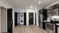 This single-floor space showcases a clean, contemporary design with light hardwood-style flooring and a neutral gray-and-white color palette that creates a bright, cohesive atmosphere throughout. The kitchen is outfitted with dark espresso cabinetry, stainless steel appliances including a full-size refrigerator and range, and light stone countertops, offering a sleek contrast that anchors the space. Track lighting along the ceiling supplements recessed lighting, ensuring the area is well-lit. A standout functional feature is the dedicated in-unit laundry closet, which houses a stacked washer and dryer unit behind dark panel doors — a practical and space-efficient solution. A second dark-finished door nearby likely leads to additional storage or a utility area. The entryway door is equipped with a modern keypad entry system, adding a layer of security and convenience. The open layout between the kitchen and laundry area promotes ease of movement, while the consistent flooring and clean wall finishes give the space a polished, move-in-ready feel suited for everyday living.