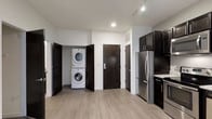 This single-floor space showcases a clean, contemporary design with light hardwood-style flooring and a neutral gray-and-white color palette that creates a bright, cohesive atmosphere throughout. The kitchen is outfitted with dark espresso cabinetry, stainless steel appliances including a full-size refrigerator and range, and light stone countertops, offering a sleek contrast that anchors the space. Track lighting along the ceiling supplements recessed lighting, ensuring the area is well-lit. A standout functional feature is the dedicated in-unit laundry closet, which houses a stacked washer and dryer unit behind dark panel doors — a practical and space-efficient solution. A second dark-finished door nearby likely leads to additional storage or a utility area. The entryway door is equipped with a modern keypad entry system, adding a layer of security and convenience. The open layout between the kitchen and laundry area promotes ease of movement, while the consistent flooring and clean wall finishes give the space a polished, move-in-ready feel suited for everyday living.