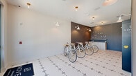 Located at Novel Laurel Sound in Sanford, Florida, this thoughtfully designed bike lounge offers a compact yet functional amenity space across a single floor. The room is defined by a striking patterned mosaic tile floor — a geometric design in white and dark accents — that lends a polished, boutique feel to the space. A fleet of white cruiser bicycles with leather saddles and wooden crate accessories are neatly arranged and available for use, serving as both a practical amenity and a visual centerpiece. The walls are finished in a two-tone palette, with crisp white on one side and a moody charcoal-gray accent wall on the other, creating a modern, curated atmosphere. White gooseneck barn-style sconces provide warm, focused lighting throughout. The accent wall features a casual photo display strung with string lights, adding a personal, community-oriented touch, while built-in cabinetry with a light wood countertop offers practical storage and workspace. A teal-framed glass entry door and large windows on one end flood the space with natural light and frame views of lush palm-lined greenery outside. A secondary area provides secure bicycle storage with glass-panel enclosures. The overall design is clean, inviting, and well-appointed.
