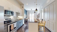This single-story residence offers a thoughtfully designed open-concept layout where the kitchen and living areas flow seamlessly together. The kitchen is a standout feature, showcasing two-tone cabinetry with white upper cabinets and slate-blue lower cabinets, dark granite countertops, a classic subway tile backsplash, and a full suite of stainless steel appliances. A central island with pendant lighting in matte black and brass provides bar seating, making it an ideal space for dining or entertaining.

The living area extends naturally from the kitchen, anchored by rich hardwood-style flooring and a bold teal accent wall. Large French doors flood the space with natural light and offer outdoor views, while a ceiling fan ensures comfort year-round. The room accommodates a comfortable seating arrangement with ample space to relax.

Both bedrooms feature plush carpeting, generous windows with plantation-style blinds, and elevated ceiling heights that enhance the sense of openness. Each room receives abundant natural light.

The bathroom is notably spacious, featuring a dual-sink vanity with granite countertops, white cabinetry, large mirrors, wood-look flooring, and a separate tiled tub-shower combination. Throughout the residence, neutral tones and modern finishes create a cohesive, contemporary aesthetic.