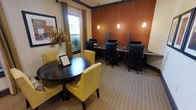 This functional and stylishly designed business center offers a versatile environment suited for both focused work and informal meetings. The space is anchored by a row of individual computer workstations along the back wall, each equipped with a monitor, privacy dividers, and ergonomic seating — ideal for residents or guests needing dedicated workspace. Pendant lights hang above the workstations, casting warm, focused illumination against a rich terracotta accent wall that adds a sense of warmth and character to the room. A casual meeting area occupies the foreground, featuring a round dark wood table surrounded by plush yellow upholstered chairs, creating an inviting spot for collaboration or informal gatherings. Natural light filters into the space through a large window dressed with elegant curtain panels, further brightening the room. A decorative floral arrangement adds a welcoming, refined touch. The flooring features a neutral-toned patterned carpet that ties the dual-purpose layout together cohesively. Crown molding along the ceiling lends a polished, finished aesthetic throughout. Overall, the space balances professional functionality with thoughtful interior design, making it an accommodating and attractive amenity.