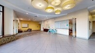 The lobby of Vista Apartment Homes, located at 2751 N 47th St in Philadelphia, PA, offers a welcoming and professionally designed space that serves as the central hub for residents and visitors alike. The layout is generous and open, anchored by a striking circular entryway framed by floor-to-ceiling windows that flood the area with natural light and offer views of the surrounding greenery. Large-format tile flooring in the entry transitions to patterned carpet throughout the interior, adding visual interest and warmth to the space. A distinctive coffered ceiling detail is adorned with oversized drum pendant lights in a yellow-gold tone, lending an elegant yet contemporary feel. The reception area features a dark wood desk prominently displaying the Vista Apartment Homes logo, creating a polished focal point. Adjacent to the reception is a comfortable lounge area furnished with upholstered sofas, patterned accent chairs, and wooden side tables, accented by bold abstract artwork in coordinating yellow and grey tones. A glass-enclosed leasing office provides a professional meeting space with ample visibility. Bright accent walls in yellow complement the neutral palette throughout, while recessed lighting and decorative pendants ensure the space remains well-lit and inviting at all times. Two bicycles near the reception desk hint at available resident amenities.