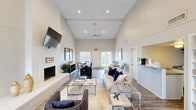 Located at 2010 S Clack St in Abilene, TX, this single-story clubhouse offers a well-appointed community gathering space designed with a modern farmhouse aesthetic. The main lounge area immediately impresses with its dramatic vaulted ceilings accented by exposed beams and a large ceiling fan, creating an open and airy atmosphere. A striking white-painted brick fireplace serves as the focal point of the lounge, topped by a wall-mounted flat-screen television, and flanked by shiplap wall paneling. The seating area is furnished with a comfortable sofa adorned with navy blue accent pillows, complementary armchairs, and glass-top coffee tables arranged atop a textured area rug, all set on warm wood-look flooring. A dedicated workstation lines one wall, featuring a long navy desk with computer monitors and industrial-style bar stools, ideal for residents needing a productive workspace. A built-in bench with decorative cushions and curated artwork adds a cozy reading nook. French doors at the rear of the space invite natural light while providing outdoor access. A separate kitchenette area includes granite countertops, white cabinetry, a microwave, and a refrigerator, offering convenient amenities for gatherings. Throughout, the neutral white and grey palette combined with navy blue accents creates a cohesive, inviting ambiance.