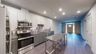 This thoughtfully designed one-bedroom, one-bathroom unit offers a practical and modern living environment across a single floor. The open-concept kitchen and living area serves as the heart of the space, anchored by a galley-style kitchen outfitted with stainless steel appliances, including a range, microwave, and dishwasher. Two-tone cabinetry pairs white upper shaker-style cabinets with gray lower cabinets, complemented by granite countertops and a decorative tile backsplash featuring a bold geometric pattern. A freestanding granite-topped island provides additional prep and dining space. Dark hardwood-style flooring runs throughout the living area, which receives natural light through a front entry door flanked by sidelights and a transom window that frames outdoor views.

The bedroom offers a comfortable footprint with plush carpeting, a large window bringing in natural light, and access to a dedicated closet. The bathroom features a granite-topped vanity with gray cabinetry, a walk-in shower with black-framed glass doors, patterned tile flooring, and recessed lighting, creating a clean, contemporary aesthetic throughout.