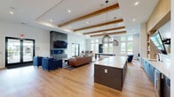 Located at 1 Haddon Blvd in Mount Laurel, NJ, this thoughtfully designed single-floor amenity space offers a generous and versatile layout for gathering, relaxing, and connecting. The main living area is anchored by a striking three-sided concrete fireplace with a wall-mounted television above, set against warm hardwood flooring and crisp white walls that create a clean, contemporary aesthetic. Exposed wood beam accents add architectural warmth overhead.

A welcoming lounge area features floor-to-ceiling windows that flood the space with natural light and offer views of surrounding greenery. Deep blue upholstered seating, including a long channeled sofa and rounded accent chairs, lends a bold, cohesive color story throughout. A kitchen bar with a white quartz countertop, wood cabinetry, beverage refrigerator, and woven pendant lighting provides an ideal social hub.

The adjacent dining and lounge area transitions seamlessly into a dedicated "Connect" room, accessible through sleek barn-style glass-paned doors. This enclosed space features rich blue walls adorned with curated artwork and sculptural wall décor, complemented by a plush area rug. Intimate booth seating with live-edge wood tables and globe pendant lights offer semi-private nooks for small group gatherings or focused work. Together, these distinct zones create a vibrant, community-centered environment.