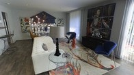 This two-floor space makes a striking impression through its bold, eclectic interior design philosophy. The living area immediately draws the eye with a tufted white sofa serving as an anchor piece, complemented by navy blue accent chairs and a warm mustard yellow chair that together create a rich, dynamic color palette. A dramatic floor lamp with a sculptural black base adds vertical interest, while a wire accent table and acrylic elements introduce varied textures and materials throughout the space. The flooring is a rich dark hardwood that grounds the room's vibrant energy. A large, vividly patterned area rug featuring warm coral, orange, and earth tones acts as a central design statement, tying together the room's diverse elements. The walls serve as a gallery, showcasing a prominent multi-panel portrait artwork alongside eclectic decorative pieces, including an architectural pediment-style wall accent and a mirrored display unit. Sculptural decorative objects, including a bold red ceramic figure, are artfully placed throughout. Sheer floor-to-ceiling curtains allow soft natural light to filter in, softening the room's bold aesthetic. The overall ambiance is that of a thoughtfully curated, artistic space that balances maximalist design with purposeful arrangement across both floors.