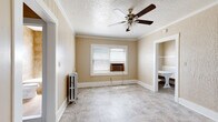 This single-floor space offers a cohesive and functional layout finished with warm neutral tones throughout. The main living area features vertical wood-paneled walls painted in a soft beige, light marble-style tile flooring, and crown molding that frames the textured ceiling above. A classic ceiling fan anchors the room, while a vintage cast-iron radiator adds a charming, utilitarian character. A window unit provides supplemental climate control, and natural light filters in through blinds-fitted windows.

An adjacent room mirrors the same warm palette and tile flooring, with a ceiling fan overhead and a window for added ventilation and light. Built-in open shelving in a nook provides convenient display or storage space. The walk-in closet is well-organized, featuring double hanging rods and a built-in white storage cabinet, offering ample room for organization.

The kitchen is efficiently designed with white cabinetry extending to the ceiling, a gas range, a granite-topped counter with an undermount sink, and tile flooring consistent with the rest of the space. A glass-paned door adds a classic architectural touch.

The bathroom is appointed with warm earth-toned tile surrounding a full soaking tub with an overhead shower, complemented by a white vanity and a natural light-admitting window, creating a clean and functional space.