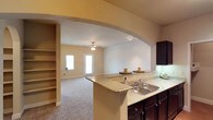 Located at 1300 Placid Circle in Waco, Texas, this single-floor residence offers a thoughtfully designed open-concept layout that maximizes both functionality and comfort. The kitchen is a standout feature, appointed with rich dark cabinetry, a granite-style countertop, a double-basin sink, and track lighting that illuminates the workspace effectively. A generous breakfast bar extends along the kitchen's edge, providing additional prep space and casual seating. A striking arched passthrough connects the kitchen to the main living area, creating a sense of openness and architectural interest. Built-in shelving is integrated into the wall adjacent to the arch, offering convenient display and storage space. The living area is carpeted and bathed in natural light from a large window and a front entry door with glass panels, contributing to a bright and welcoming atmosphere. A ceiling fan with a light fixture is centrally positioned in the living room, ensuring year-round comfort. The overall color palette features warm neutral tones on the walls and ceilings, complementing the darker cabinetry and creating a cohesive, inviting interior throughout the single-level floor plan.