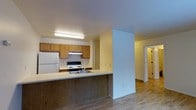 This single-floor space is designed with functional simplicity and an open, connected layout. The kitchen serves as a central feature, outfitted with warm honey-toned wood cabinetry that extends to the upper and lower sections of the walls, providing ample storage. A freestanding electric range with an overhead exhaust hood, a full-size refrigerator, and a sink integrated into the peninsula counter complete the kitchen's practical setup. The peninsula extends outward, creating a natural divider between the kitchen and the adjoining living area while maintaining an open sightline throughout the space. Light-toned hardwood flooring runs continuously through the kitchen, living area, and hallway, lending a cohesive and airy feel to the interior. The walls are finished in a clean, neutral white, complementing the warm wood tones and contributing to a bright, welcoming atmosphere. A wide hallway visible from the main area leads to additional rooms, with interior doors and recessed lighting adding to the sense of depth and accessibility. Overhead fluorescent lighting in the kitchen ensures a well-lit workspace. The overall layout balances openness with defined zones, making the space both practical and comfortable.