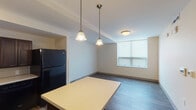 This single-floor residential unit offers a well-designed, functional layout with a clean, modern aesthetic. The open-concept kitchen is a central highlight, featuring rich dark-stained cabinetry with ample upper and lower storage, light countertops, a full suite of black appliances including a refrigerator, dishwasher, and range, and durable hardwood-style flooring. A kitchen island with a light countertop surface extends into the dining area, which is illuminated by two elegant pendant lights and benefits from natural light through a frosted window. The bedrooms are bright and generously proportioned, each finished with the same hardwood-style flooring, neutral warm-toned walls, and windows fitted with roller blinds that allow for controlled natural light. The bathroom is thoughtfully designed with accessibility in mind, featuring strategically placed grab bars near the toilet and along the walls, a wide-format vanity with a white countertop and dark wood base, large-format tile flooring, and warm ambient lighting above the mirror. Throughout the unit, the consistent use of neutral tones, clean lines, and durable finishes creates a cohesive and practical living environment suited for a variety of needs.