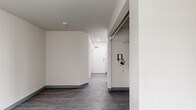 This compact single-floor studio unit offers a straightforward, functional layout designed for efficient use of space. The main living area features neutral beige walls, grey wood-look vinyl flooring, and dark baseboards that provide a clean, modern contrast throughout. A tall, narrow window fitted with horizontal blinds allows natural light to filter into the room, complemented by a wall-mounted panel radiator that serves as the primary heating source. The unit includes a dedicated closet area equipped with wire shelving and hanging rods, providing practical storage solutions. Adjacent to the entry, a full-size refrigerator is conveniently positioned near the front door, which features a keypad entry system. The corridor-style entryway leads seamlessly into the living space, maintaining an open and uncluttered flow. The bathroom is outfitted with a pedestal sink, toilet, and a full alcove bathtub with a shower, all finished in white with grey vinyl flooring consistent with the rest of the unit. Smoke detectors are installed throughout for safety. The overall aesthetic is minimalist and clean, with a neutral color palette that offers a blank canvas for personalization.