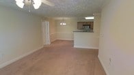 This single-floor space showcases an open-concept layout that seamlessly connects the living, dining, and kitchen areas, creating a spacious and functional environment. The living and dining areas are finished with warm, neutral-toned wall-to-wall carpeting that adds comfort underfoot, while the textured ceiling contributes a classic architectural detail throughout. A ceiling fan with a light fixture serves the main living area, and an elegant chandelier marks the dining space, providing both ambient and focused lighting. The kitchen is positioned at the far end of the open layout, anchored by a peninsula-style breakfast bar with a granite-look countertop that provides a natural visual divider between the cooking and living zones. The kitchen features medium-toned wood cabinetry, a stainless steel microwave, and under-cabinet lighting that illuminates the workspace. Soft, neutral tones on the walls throughout the unit lend a clean and welcoming ambiance. A paneled door provides access to an adjacent room or utility space, adding to the functional versatility of the floor plan. Overall, the space offers an inviting, easy-flowing layout well-suited for everyday living and entertaining.