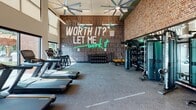 This single-floor fitness center in Seneca, SC is designed to inspire and accommodate a full range of workout styles. The space is architecturally distinguished by soaring ceilings with clerestory windows that flood the interior with natural light, complemented by oversized industrial ceiling fans that keep the environment comfortable during intense sessions. Large windows along the perimeter offer views of the surrounding area, reinforcing an open, energizing atmosphere.

The cardio zone features a row of commercial-grade treadmills positioned to take advantage of the natural light and outdoor sightlines. Adjacent to this area, a dedicated free weights section includes a comprehensive dumbbell rack with a full range of weights and an adjustable bench, set against a dramatic accent wall. That wall serves as the focal point of the gym — a bold, typographic mural in dark tones punctuated by a vibrant teal neon sign reading "Is it worth it? Let me work it," delivering clear motivational energy to the space.

The strength and functional training area is anchored by cable machine systems and a mirrored wall that visually expands the space. A heavy punching bag with TRX suspension straps, kettle bells, and medicine balls round out the functional fitness offerings. Exposed brick walls add an industrial warmth that balances the modern equipment, creating a cohesive and high-energy training environment throughout.