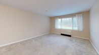 Located at 5945 Vinecroft Dr in Clarence Center, NY, this single-floor, one-bedroom, one-bathroom unit offers a practical and comfortable layout. The open-concept kitchen and living area serves as the heart of the home, where warm honey-toned cabinetry pairs with stainless steel appliances — including a refrigerator and range — and light-toned countertops. Hardwood-style flooring runs through the kitchen, transitioning to plush carpeting in the living and bedroom areas, creating a welcoming contrast of textures. The living room receives natural light through a large window overlooking a green outdoor area, with a wall-mounted heating unit adding a functional touch. The bedroom is a comfortable, well-proportioned space with a window view and access to two closets, one of which features built-in shelving for ample storage. The bathroom includes a wood-framed mirrored medicine cabinet, a single-sink oak vanity, a full soaking tub with grab bars, and tile flooring. Neutral wall tones and clean white trim throughout lend the space a bright, tidy aesthetic. A dedicated entry area with closet access adds convenience upon arrival, rounding out this well-organized and efficiently designed home.