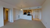 This single-floor space showcases an open-concept design that seamlessly integrates the living area and kitchen into one cohesive environment. The living space is finished with neutral carpeting, creating a warm and comfortable atmosphere, while the kitchen and entry areas transition to hardwood flooring, offering a practical and visually appealing contrast.

The kitchen is defined by a peninsula with a dark countertop, providing both workspace and a natural division between the cooking and living zones. Warm-toned wood cabinetry lines the kitchen walls, complemented by black appliances including a refrigerator and range. Overhead, a track lighting system and two pendant lights — one a classic dome style — illuminate the kitchen effectively.

The living area is generously proportioned, with smooth white walls and recessed lighting overhead contributing to a clean, airy feel. A HVAC vent is integrated into one wall, ensuring climate control throughout the space. A light-toned entry door with a standard viewer is visible, and a sliding door on the opposing side hints at additional storage or room access. The overall ambiance is practical and understated, well-suited for everyday living.