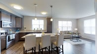 This single-floor space showcases a thoughtfully designed open-concept layout that seamlessly connects the kitchen and living areas. The kitchen features dark espresso cabinetry that contrasts elegantly with light countertops, creating a striking visual balance. A central island with a bright countertop serves as both a functional workspace and a casual dining area, accommodated by four white upholstered bar stools with dark legs. Pendant lighting in brushed nickel hangs above the island, complementing the flush-mount ceiling fixture overhead. The kitchen is equipped with stainless steel appliances and a double sink positioned beneath a window, allowing natural light to fill the space. The open living area flows naturally from the kitchen, featuring warm hardwood flooring, a neutral-toned sofa, a patterned area rug, and a glass-top coffee table. Large windows throughout the space invite generous amounts of natural light, enhancing the bright, airy atmosphere. The overall aesthetic is modern and cohesive, with a neutral color palette of whites, grays, and warm wood tones creating a welcoming and functional environment.