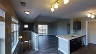 This single-floor residence showcases an open-concept layout that seamlessly integrates the kitchen and living areas into one cohesive space. Dark hardwood-style flooring runs throughout, providing a rich, contemporary contrast against the cool gray-toned walls. The kitchen features dark cabinetry paired with a bright white countertop peninsula that doubles as a breakfast bar, complete with a stainless steel sink. A tiled backsplash adds a subtle decorative element to the cooking area. The living space is bathed in natural light through a large sliding glass door that opens onto a private balcony, offering outdoor views of surrounding greenery. A staircase with a white-railed banister is visible near the entry, adding architectural interest to the layout. Overhead, a track lighting fixture in the main area and a flush-mount ceiling light provide ample illumination throughout the space. A small window near the staircase provides additional natural light. The overall ambiance is modern and well-appointed, with clean lines and a neutral color palette contributing to a sleek, urban aesthetic.