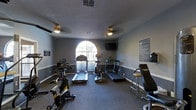 This fitness center is designed to provide a practical and comfortable workout environment. The room features gray walls accented by a white chair rail, giving the space a clean, structured appearance. Underfoot, dark carpeting covers the floor throughout, adding a degree of sound absorption suited to an active space.

The centerpiece of the room is an arched window along the back wall, which allows natural light to filter in and provides a welcoming focal point. Ceiling fans with attached light fixtures are positioned throughout, ensuring adequate ventilation and illumination during workouts. A wall-mounted flat-screen television is positioned at eye level, offering entertainment for users during cardio sessions.

The equipment lineup includes multiple treadmills, an elliptical trainer, a stationary upright bike, and a spin bike, catering to a variety of cardio preferences. A weight bench and a bar stool-style seat are situated along one side, providing space for strength training or rest. Informational signage, including fitness center rules and guidelines, is posted on the walls. The textured ceiling adds a finishing touch to this straightforward, well-equipped fitness space.