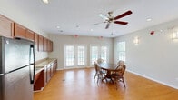 Located at 3220 Hoffmeyer Rd in Florence, South Carolina, this single-floor community space offers a well-organized, open layout that seamlessly integrates a kitchen, dining area, and living room into one cohesive environment. The interior is finished with warm light hardwood flooring throughout, complemented by neutral-toned walls that create a bright and welcoming atmosphere.

The kitchen is outfitted with rich, dark-stained wood cabinetry extending from floor to ceiling, offering generous storage capacity. Light-toned countertops run the full length of the wall, accompanied by a stainless steel sink, a built-in microwave, and a full-size refrigerator. Recessed lighting above adds functional illumination to the workspace.

The dining area features French doors with sidelight panels that open to an outdoor area with a visible pool, flooding the space with natural light. A dining table with Windsor-style chairs provides a comfortable setting for gatherings.

The living area is equally well-lit, with multiple windows fitted with white horizontal blinds. Wall-mounted sconce lighting adds a warm accent, and the room comfortably accommodates seating arrangements. An additional area features a wall-mounted television and a side entry door. Multiple exit points throughout the space enhance accessibility and flow.