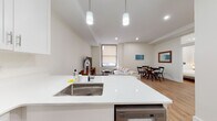 This thoughtfully designed single-floor residence offers a clean, contemporary aesthetic throughout, characterized by crisp white walls, warm hardwood flooring, and an abundance of recessed lighting. The open-concept living and dining area flows seamlessly from the kitchen, creating a spacious and inviting communal space. A large window fitted with a roller shade allows natural light to fill the living room, which features a comfortable sectional sofa, a round wood coffee table, and a dedicated dining area with a classic round table and chairs. The kitchen is well-appointed with white shaker cabinetry, stainless steel appliances including a refrigerator, range, microwave, and dishwasher, complemented by white quartz countertops and a peninsula with bar seating. An in-unit washer is conveniently situated nearby. The space includes two bedrooms, each furnished with a queen bed, upholstered headboard, and minimalist nightstands, with one bedroom offering a walk-in closet. The bathroom is a standout feature, showcasing large-format marble-style tile flooring, subway-tiled walls, and a full soaking tub with an overhead rain shower and glass enclosure. The overall ambiance is bright, clean, and modern.