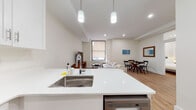This thoughtfully designed single-floor residence offers a clean, contemporary aesthetic throughout, characterized by crisp white walls, warm hardwood flooring, and an abundance of recessed lighting. The open-concept living and dining area flows seamlessly from the kitchen, creating a spacious and inviting communal space. A large window fitted with a roller shade allows natural light to fill the living room, which features a comfortable sectional sofa, a round wood coffee table, and a dedicated dining area with a classic round table and chairs. The kitchen is well-appointed with white shaker cabinetry, stainless steel appliances including a refrigerator, range, microwave, and dishwasher, complemented by white quartz countertops and a peninsula with bar seating. An in-unit washer is conveniently situated nearby. The space includes two bedrooms, each furnished with a queen bed, upholstered headboard, and minimalist nightstands, with one bedroom offering a walk-in closet. The bathroom is a standout feature, showcasing large-format marble-style tile flooring, subway-tiled walls, and a full soaking tub with an overhead rain shower and glass enclosure. The overall ambiance is bright, clean, and modern.