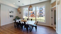 This single-floor space presents a cohesive, modern design centered around an open-concept layout that connects several functional areas. The living area serves as a welcoming focal point, anchored by a wall-mounted fireplace framed in natural wood shiplap paneling, complemented by a wall-mounted television above. Warm hardwood-style flooring runs throughout, tying each zone together seamlessly. The living area accommodates generous seating alongside a live-edge coffee table, textured area rug, and bold black-and-white framed artwork that adds visual contrast to the neutral palette.

The dining area is a standout feature, bathed in natural light from a large multi-pane window wall that frames views of an outdoor playground and green space. A sizable dining table seating eight is suspended beneath woven pendant lights, creating an inviting gathering atmosphere. An additional seating nook with rattan chairs and a floor lamp offers a more intimate setting.

The kitchen is thoughtfully appointed with two-tone cabinetry in white and natural wood, white subway tile backsplash, open floating shelves, stainless steel appliances including a full-size refrigerator and dishwasher, and a matte black faucet over an undermount sink. Recessed lighting throughout ensures the entire space remains bright and functional.