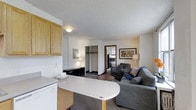 This single-floor unit presents an open-concept layout that seamlessly connects the kitchen and living areas, creating an efficient and practical living space. The kitchen is outfitted with light oak cabinetry providing ample storage, a white countertop that extends into a peninsula-style breakfast bar, and standard appliances including a dishwasher. The textured white ceiling and neutral wall tones lend a clean, understated aesthetic throughout.

The living area is carpeted and comfortably accommodates a gray upholstered sofa and armchair arranged around a round coffee table, with framed artwork adorning the walls and floor lamps providing warm ambient lighting. A recessed closet with a hanging rod offers built-in storage and is centrally visible from the main living space.

A doorway leads to a separate room beyond the living area, suggesting additional functional space such as a bedroom or den. Natural light filters in through a window visible in the background, contributing to the overall brightness of the unit. The layout is straightforward and well-suited for efficient everyday living.